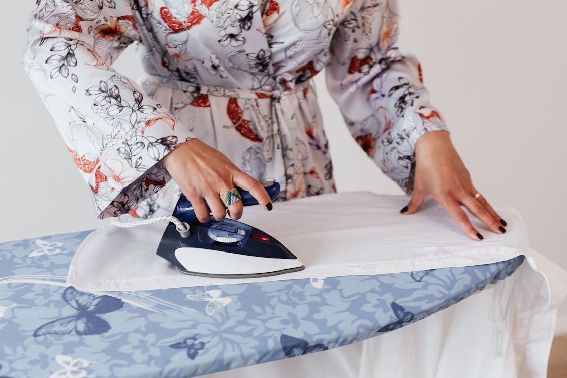 Free Crop faceless female in stylish silk robe ironing formal white shirt on blue ironing board by using contemporary small iron against white plain wall Stock Photo